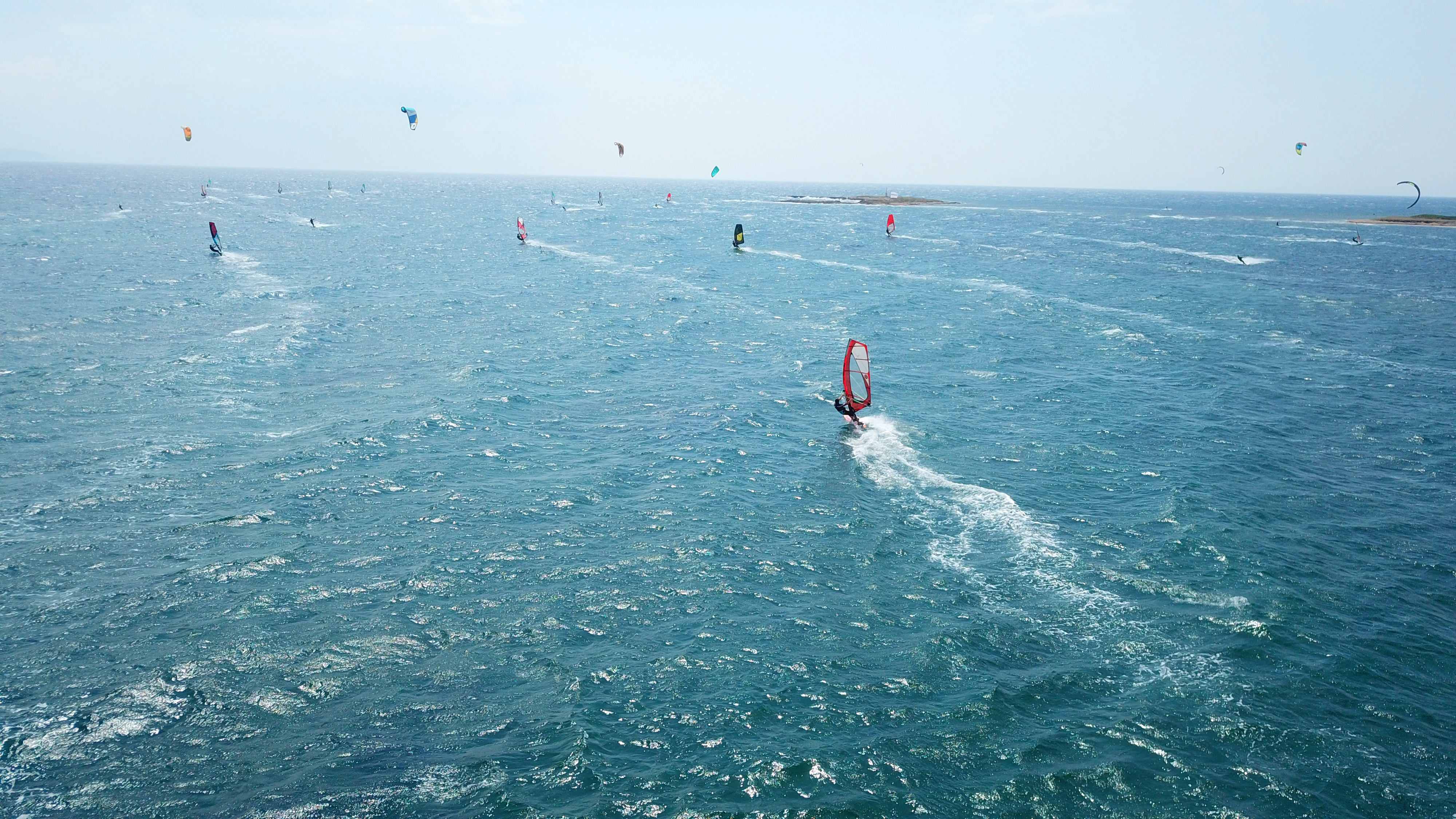 KJ Reports, Middle East — A high-angle shot of Iranian IRGC fast attack craft maneuvering in the turquoise waters of the Strait of Hormuz near a large commer…