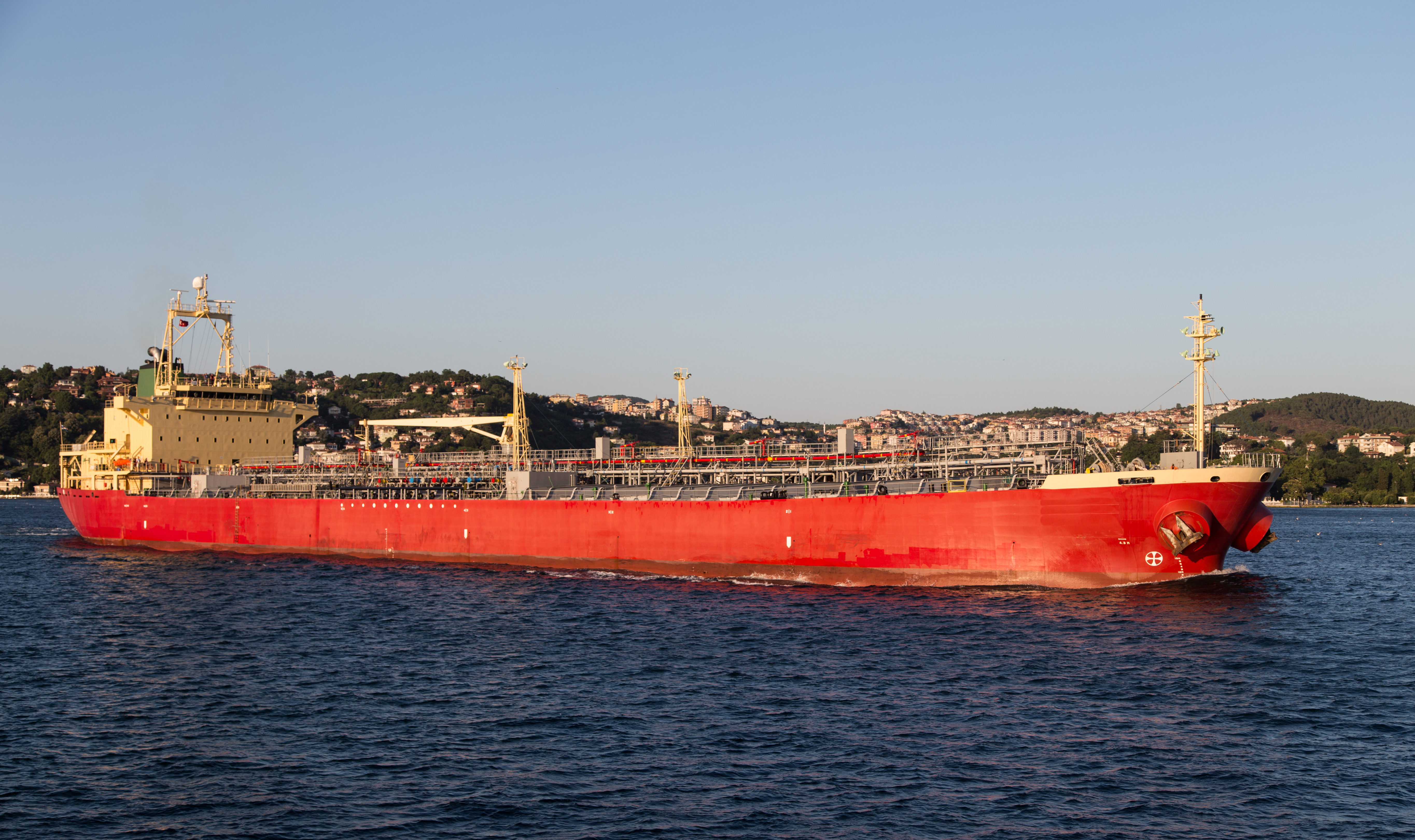 KJ Reports, Middle East — A large commercial oil tanker navigating through a narrow strait at dusk, with the blurred lights of a coastal industrial city in t…
