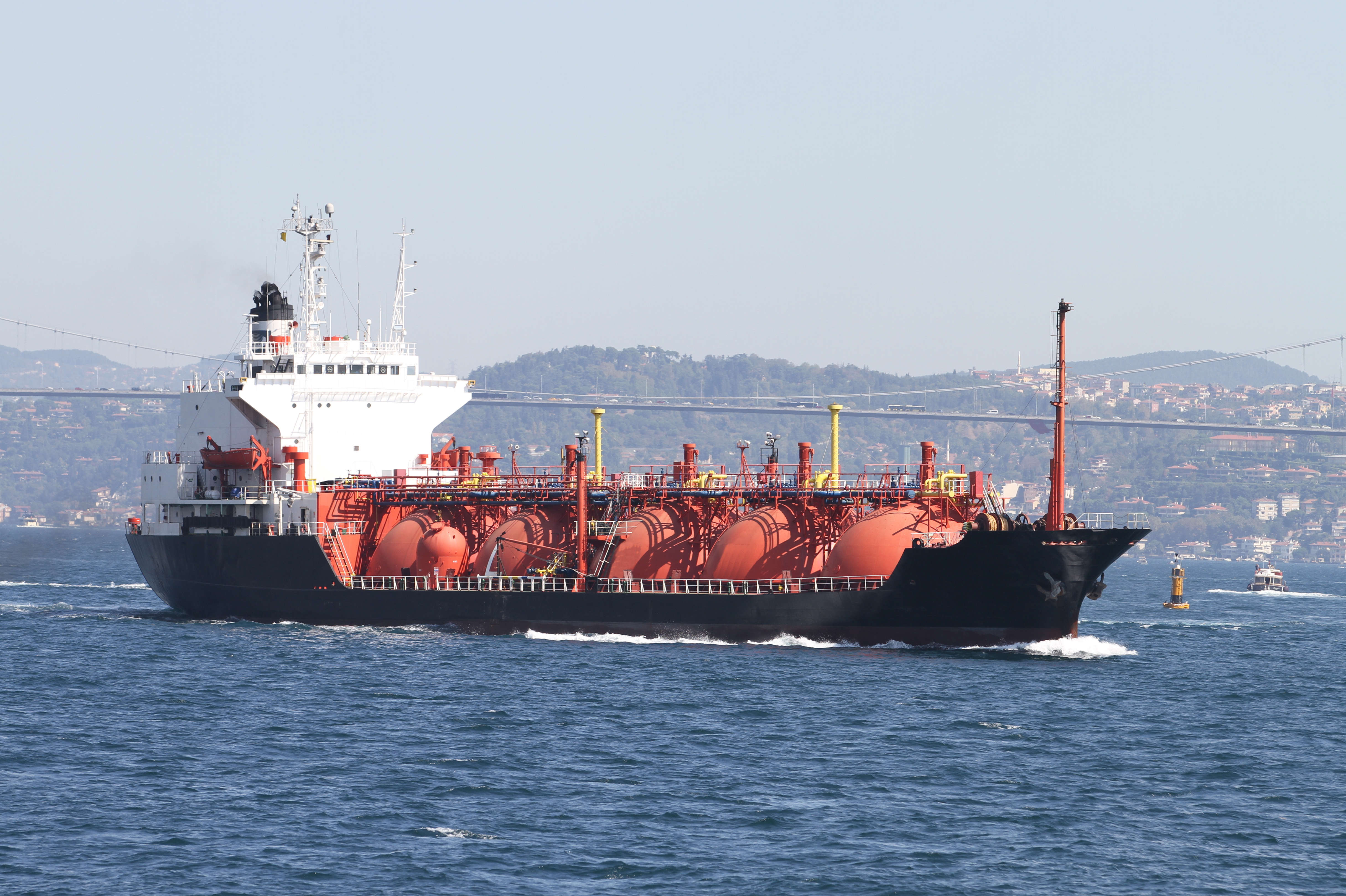 KJ Reports, Middle East — A wide-angle shot of a massive oil tanker navigating the narrow waters of the Strait of Hormuz at dusk, with the silhouette of a sm…